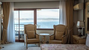 A hotel room with a bed, two armchairs, a small table, a nightstand, and a view of the ocean through large windows. - Akol Hotel (Çanakkale)