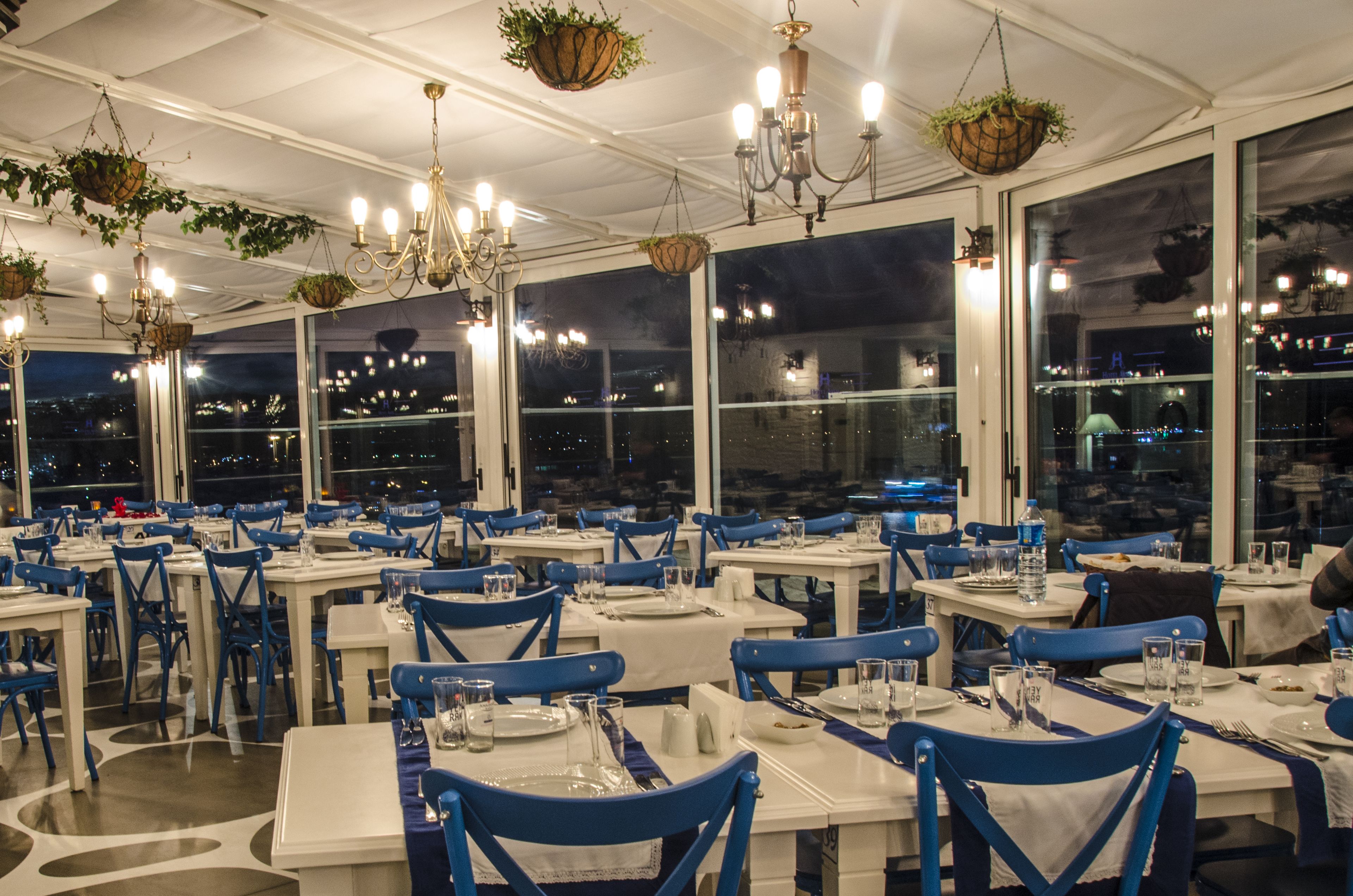 Un restaurante con varias mesas y sillas, ventanas amplias y plantas colgantes.