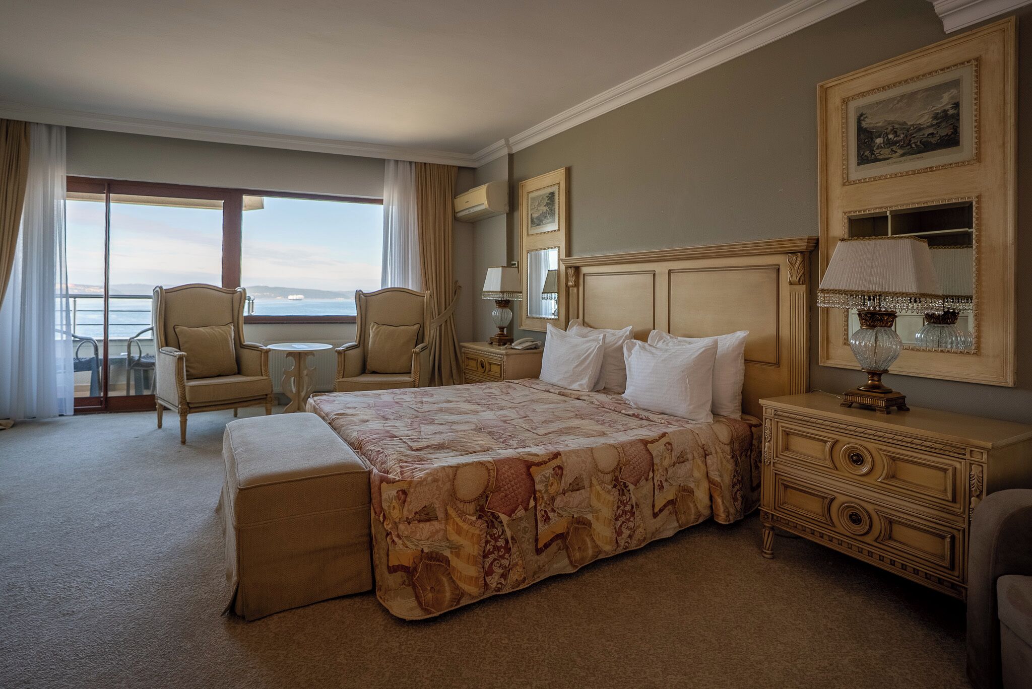 A hotel room with a large bed, a sitting area with a chair and table, a dresser, and a view of the sea through large windows.