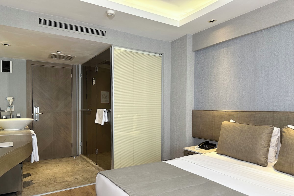 A modern hotel room with a large bed, a bedside table with a phone, a bathroom with a sink and mirror, and a walk-in closet.