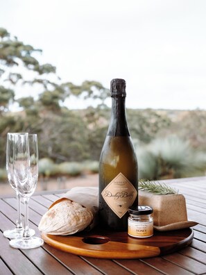 A bottle of Dudley Bubble wine, a glass, a loaf of bread, and a honey jar on a wooden table with a blurred outdoor background. - Ecopia Retreat (Seddon)