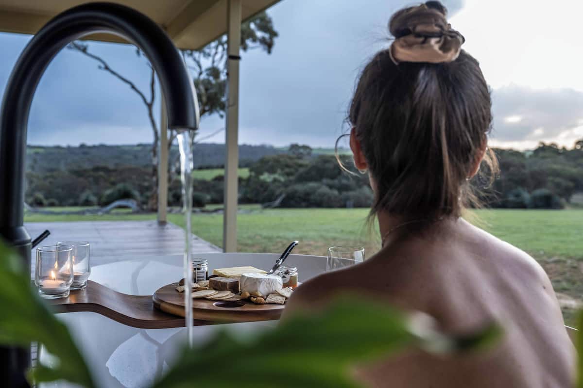 A person seated at a table with a view of a grassy landscape. The table has a wooden tray with food items and glasses.