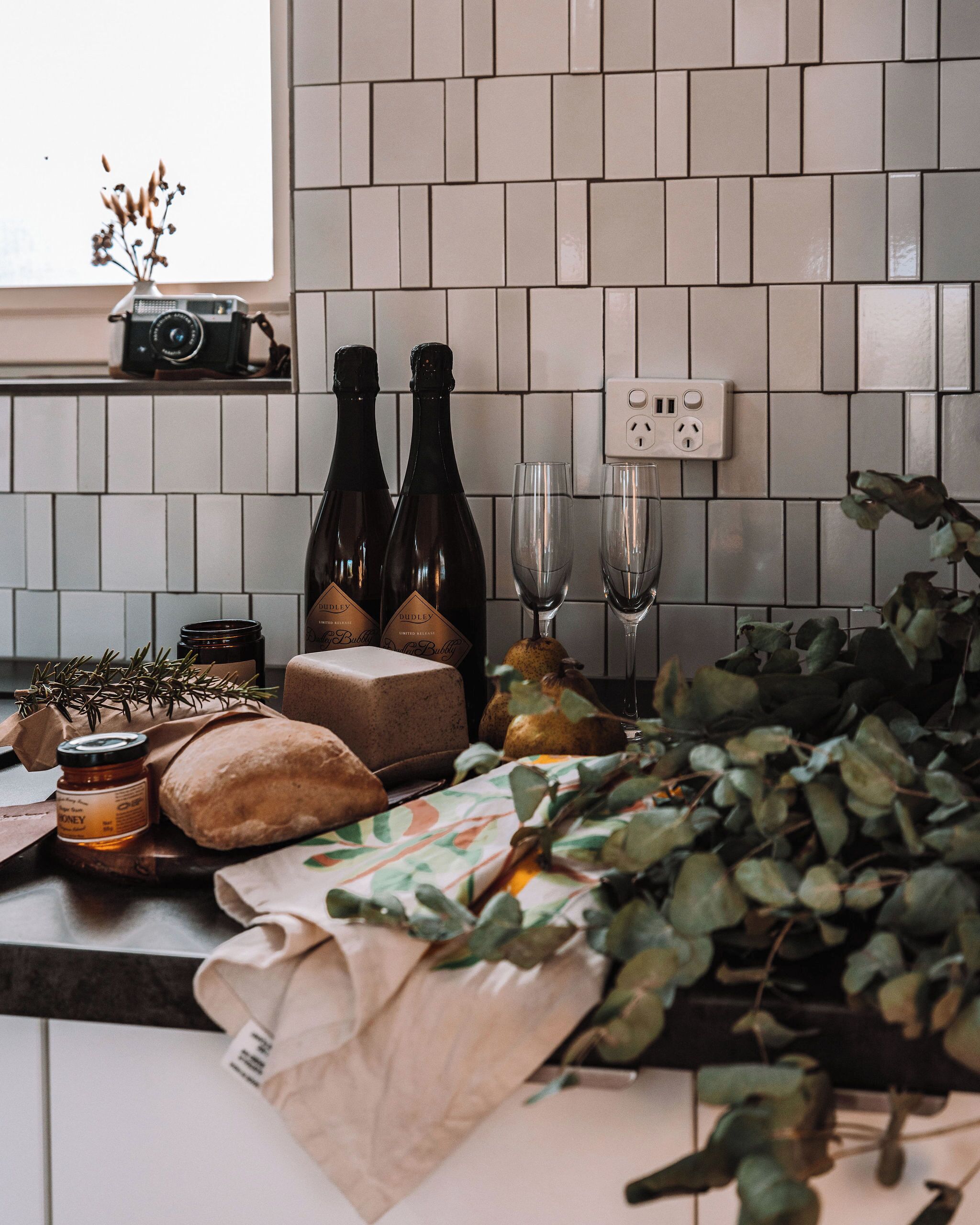 A kitchen with white tiled backsplash, two bottles of wine, a jar of honey, a loaf of bread, and a bunch of green leaves on the counter.
