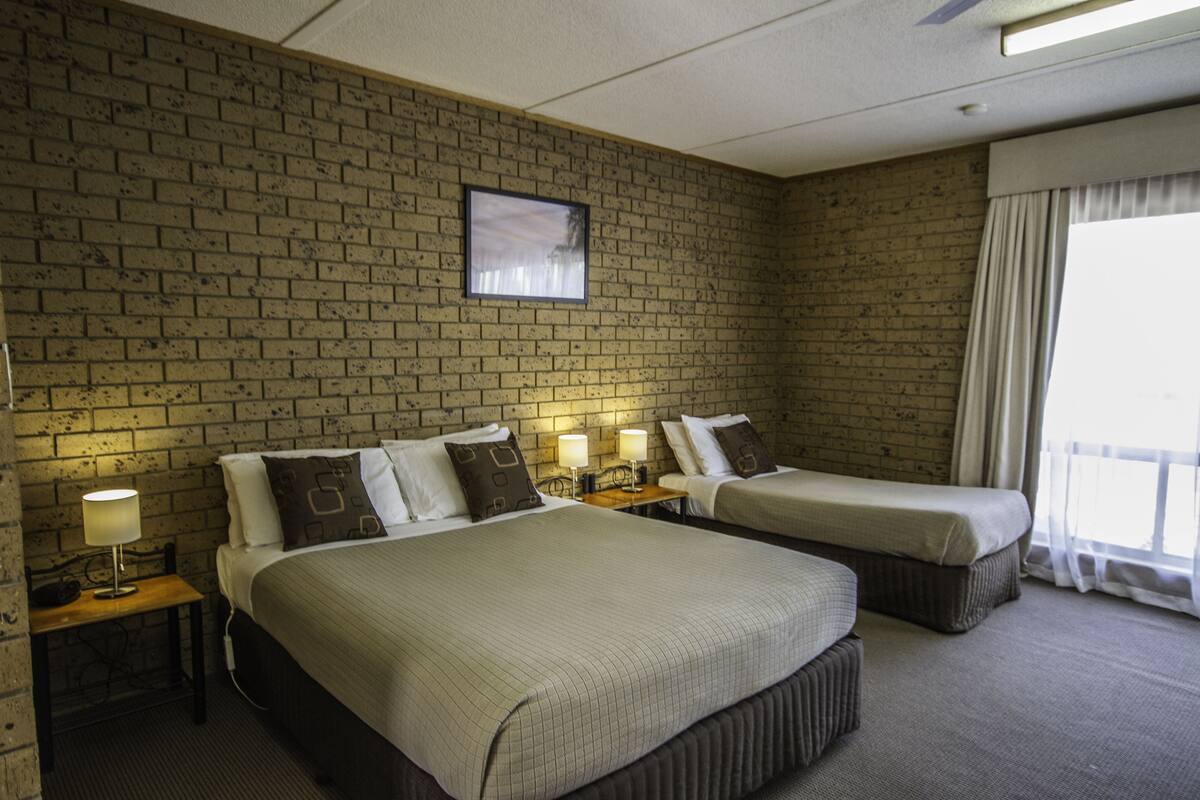 A hotel room with two beds, a brick wall, a framed picture, bedside lamps, and a window with curtains.