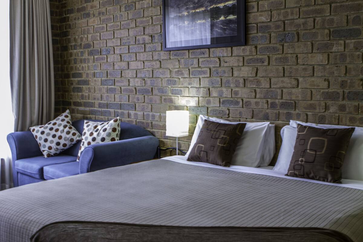 A hotel room with a brick wall, a bed with a white bedspread, a blue armchair with patterned pillows, and a bedside table with a lamp.