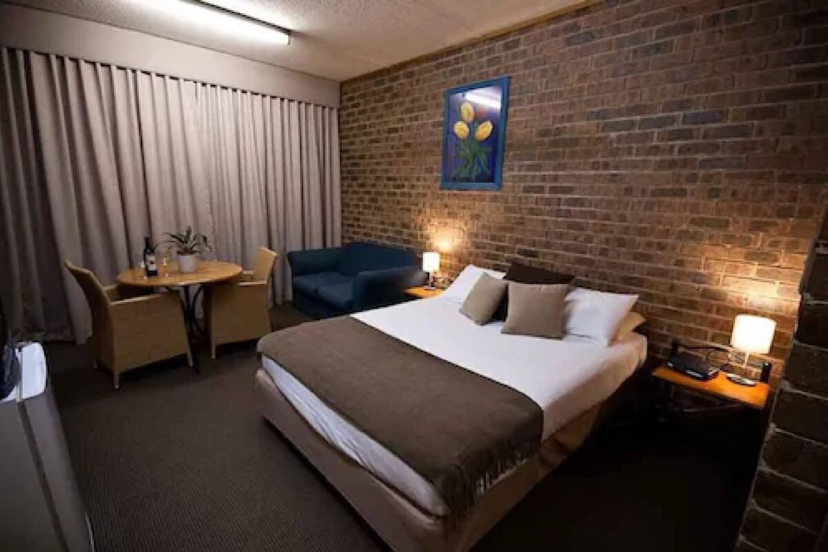 A hotel room with a bed, a desk, a chair, a small table, and a nightstand with a lamp.