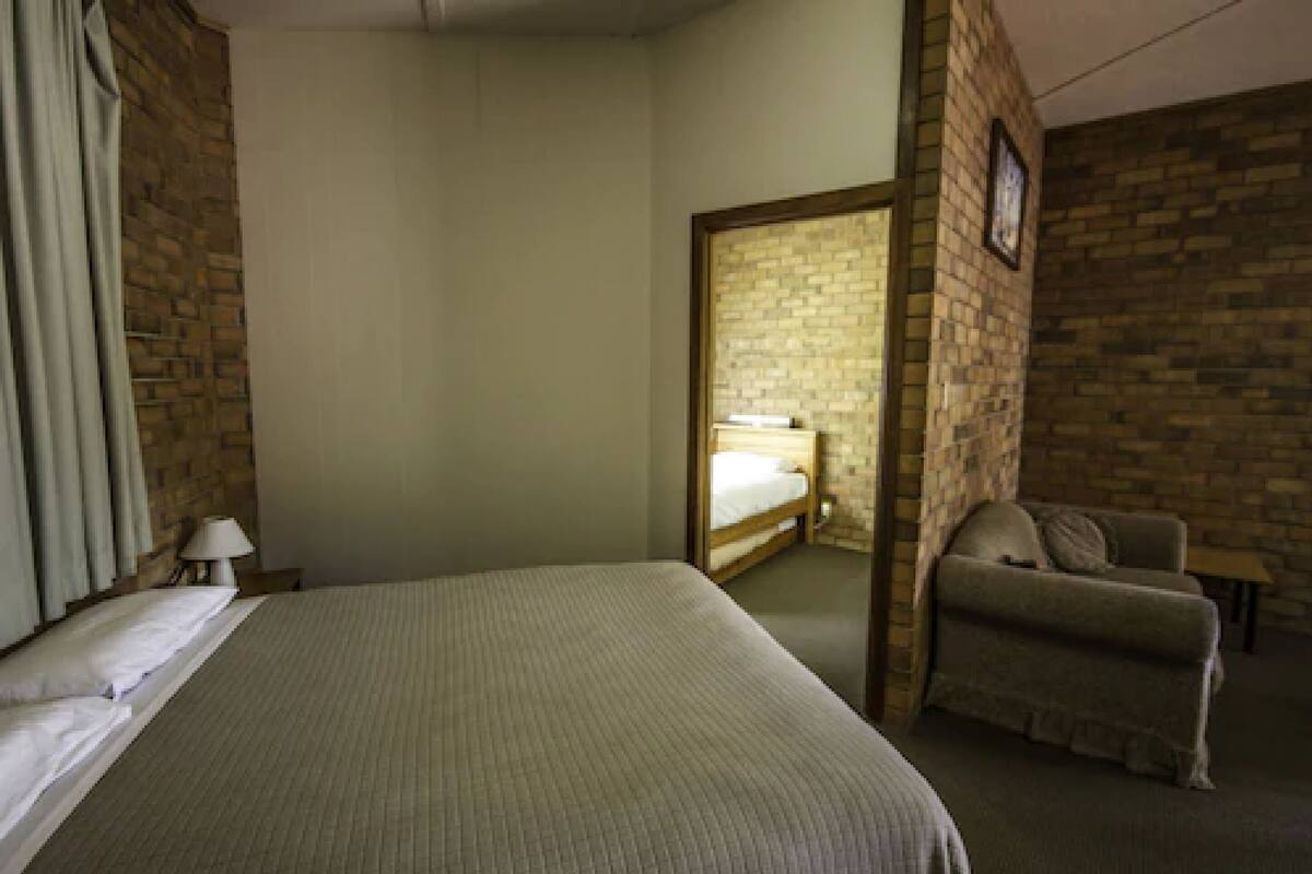 A hotel room with a bed, a chair, a lamp, and a brick wall.