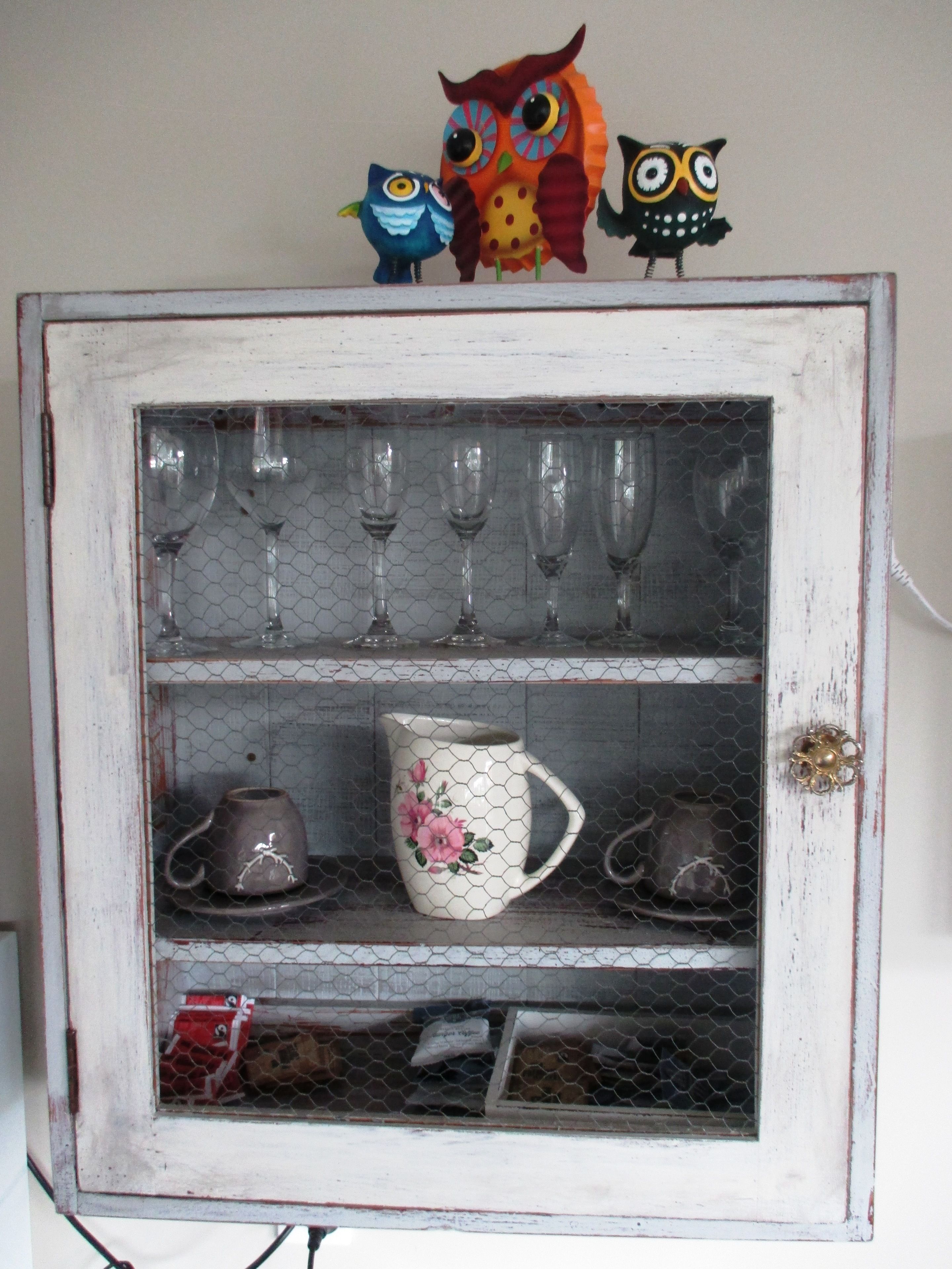 A vintage cabinet with glass doors, containing wine glasses, a floral mug, and two small figurines.