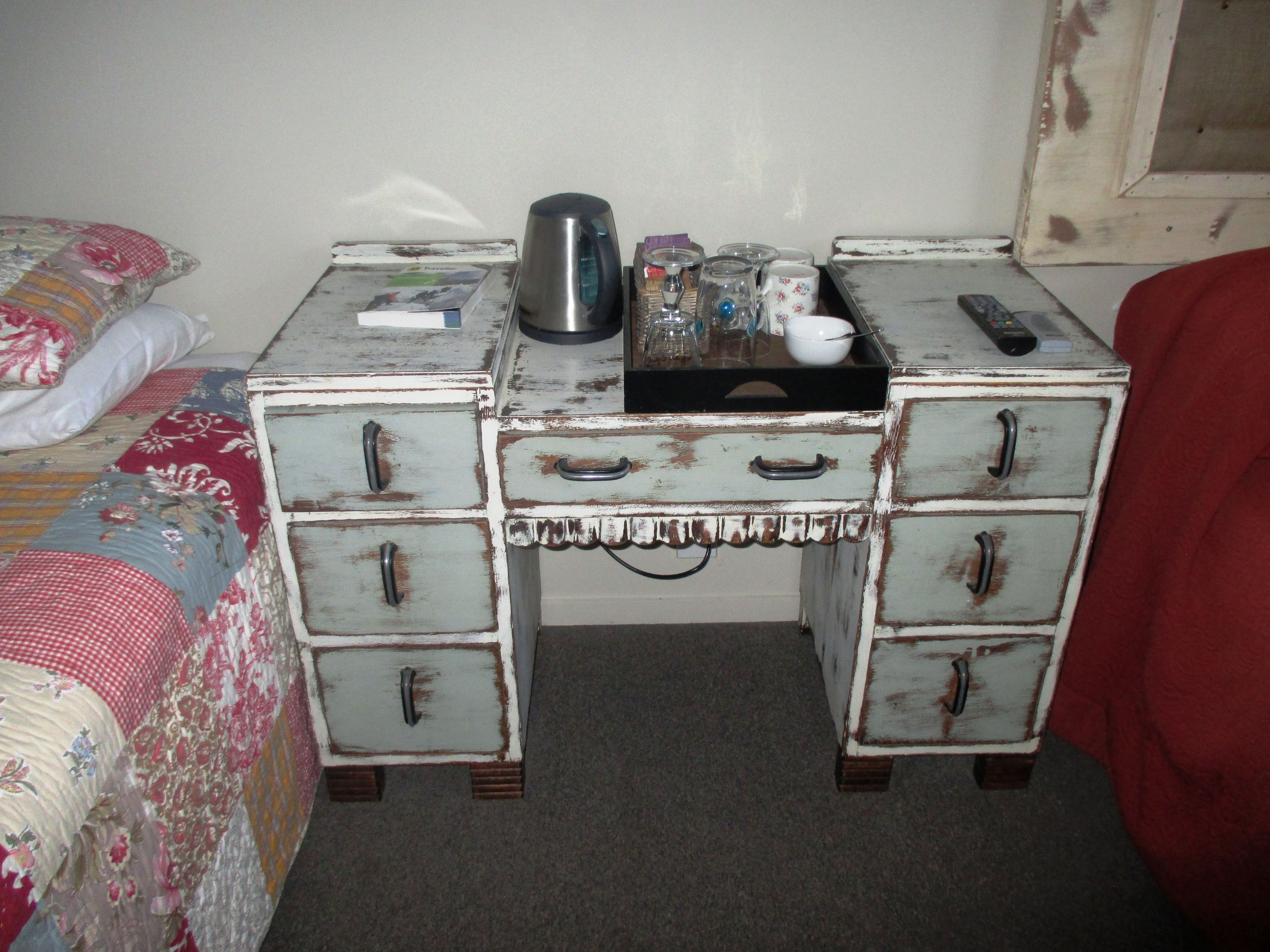 A small, rustic bedroom with a bed, a dresser with multiple drawers, and a nightstand with a kettle and other items.