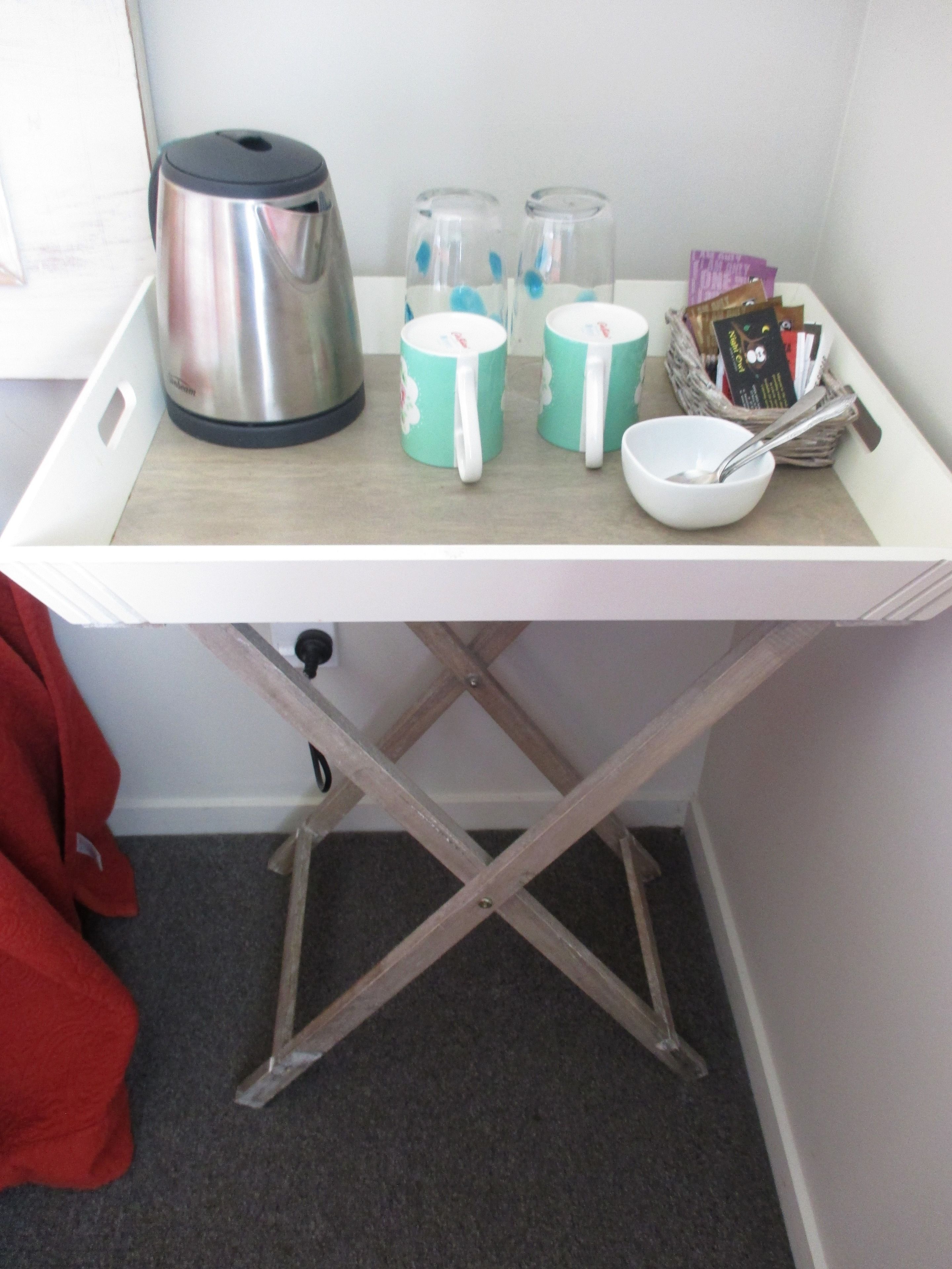 A white tray table with a stainless steel kettle, two teal mugs, a white bowl with a spoon, and a basket of cards.