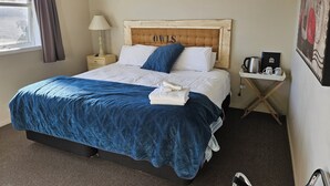 A neatly made bed with a blue quilt, a wooden headboard with the word "OWLS" on it, a bedside table with a lamp, and a small tray with coffee items. - Fat Owl Motel, Bar & Eatery (Piopio)
