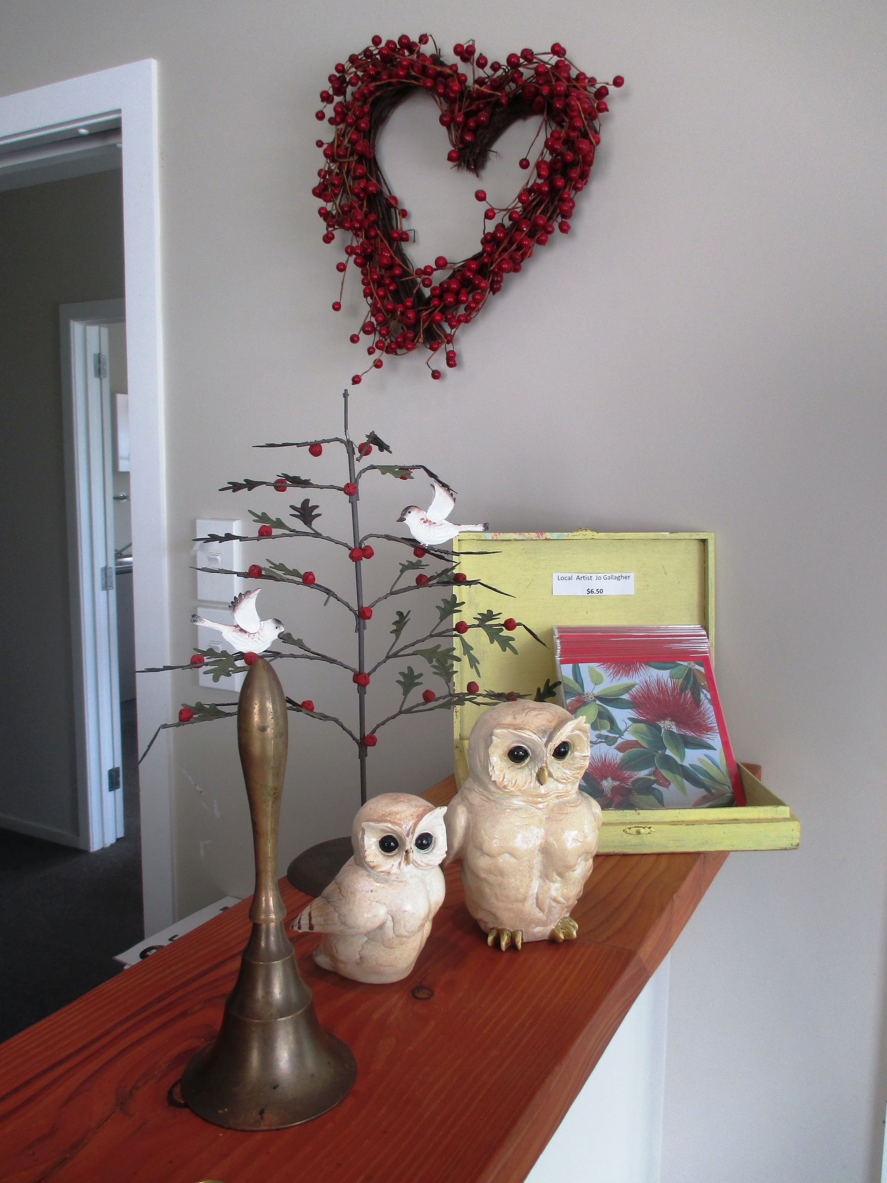 A wooden shelf with a decorative tree, two owl figurines, a brass candle holder, and a heart-shaped decoration with red berries.