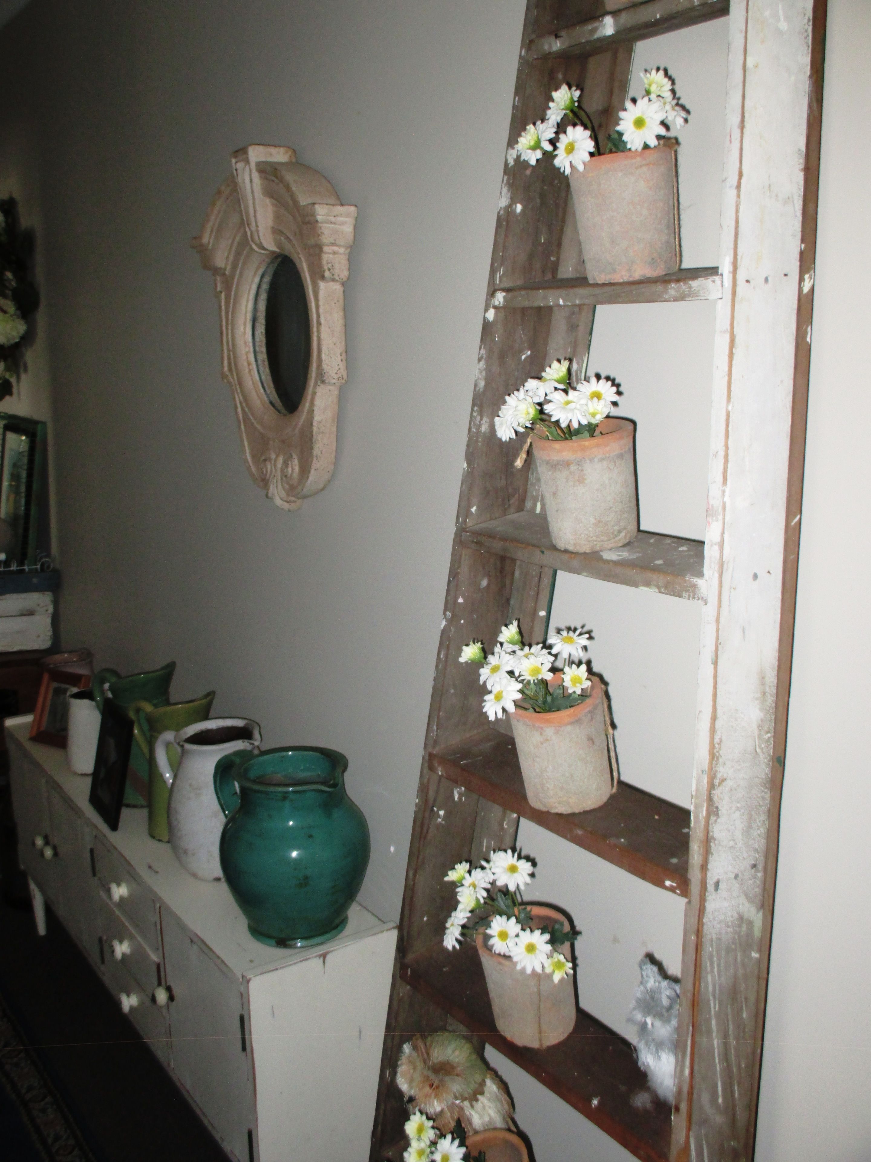 A rustic interior with a wooden ladder shelf holding potted flowers, a stone wall-mounted mirror, and a variety of ceramic containers on a white dresser.