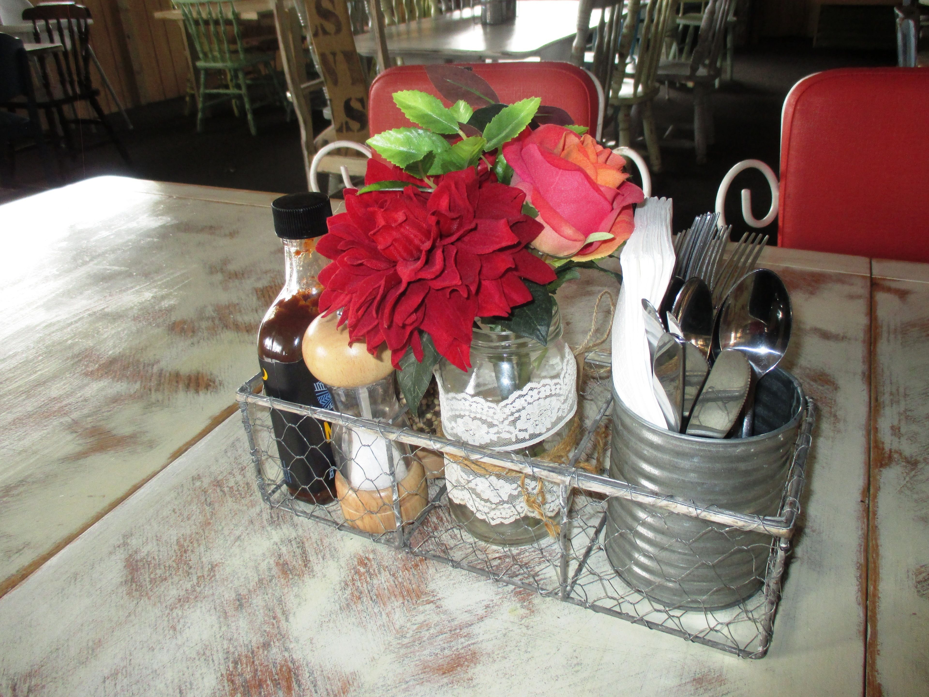 A table with a metal tray holding a bottle, a jar, a lace doily, and a metal cup with utensils.