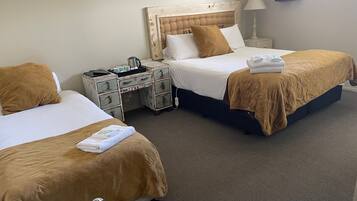 Habitación de hotel con dos camas, cada una con ropa de cama marrón y almohadas blancas. Pequeño mesita de noche con lámpara y mesita con lámpara. El cuarto tiene un diseño sencillo y limpio con piso alfombrado.