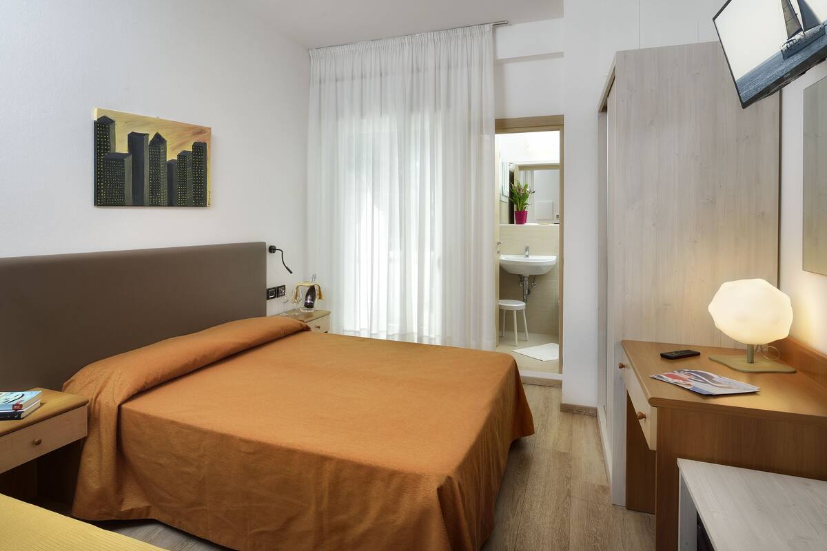 A hotel room with a bed, bedside table, lamp, and a painting of a cityscape.