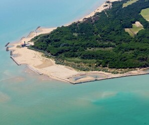 An aerial view of a coastal area with a sandy beach, dense green forest, and clear turquoise waters. - Hotel Golf (San Michele al Tagliamento)