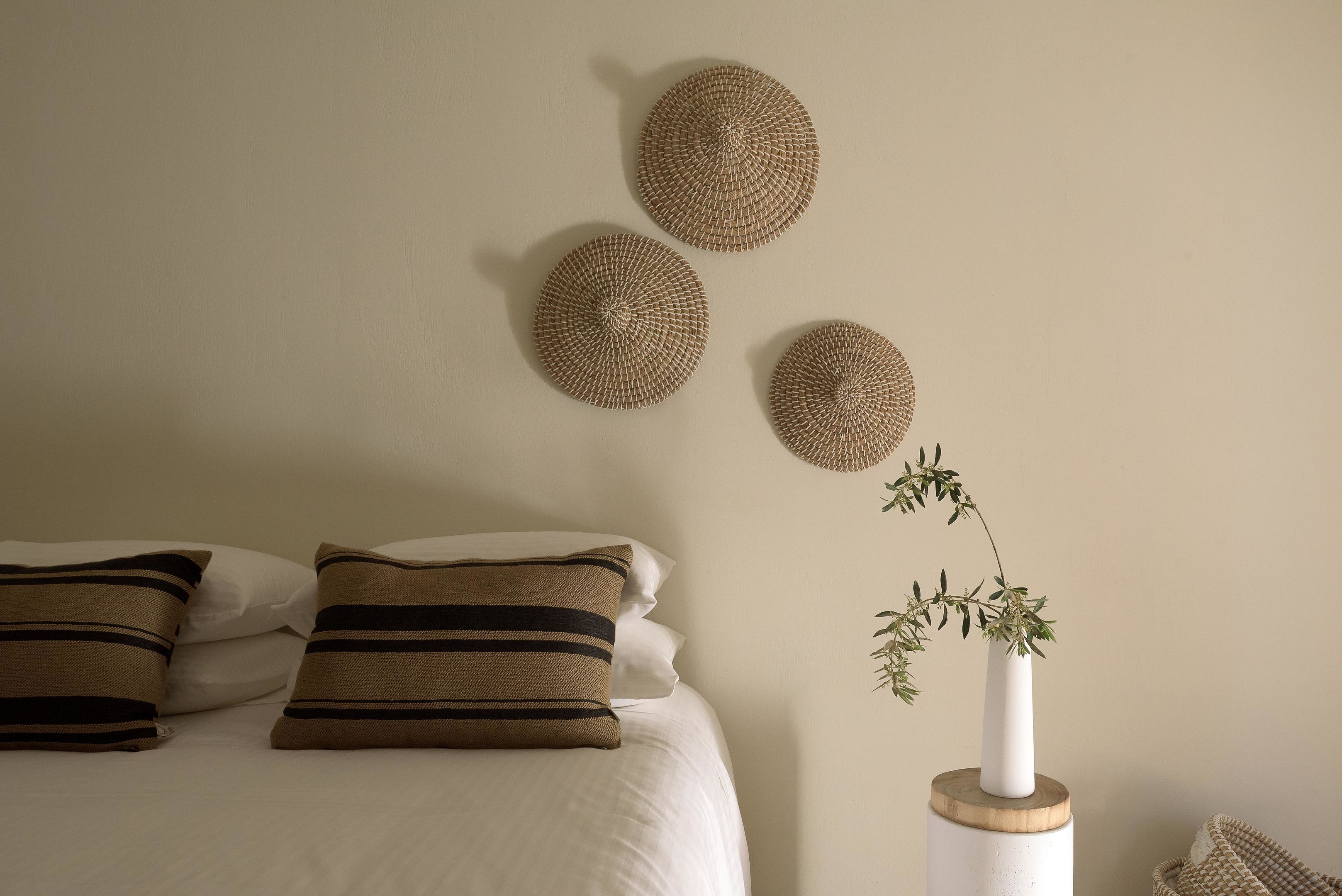 Una cama con almohadillas a rayas, un jarrón blanco con una planta y tres adornos de pared tejidos.