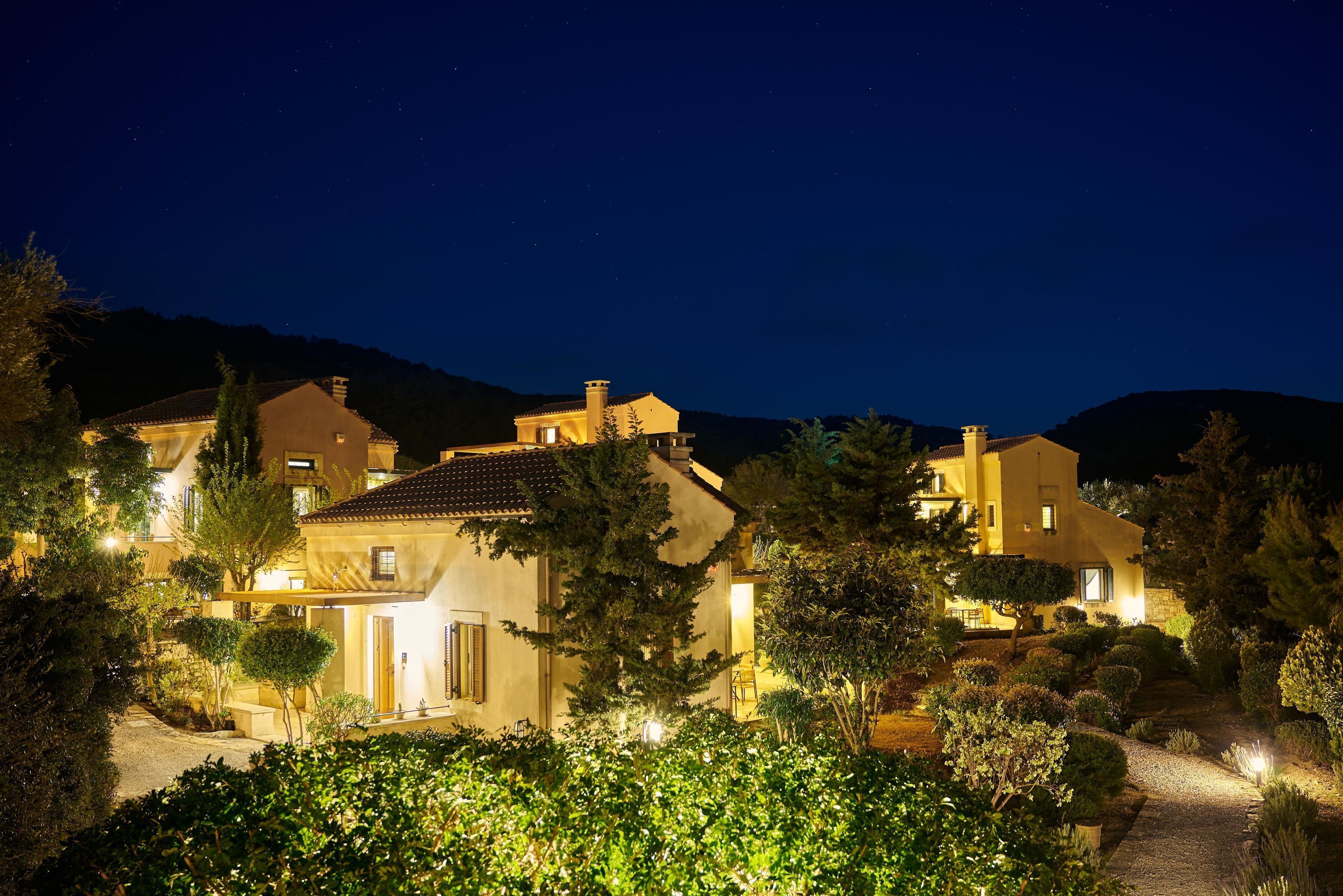 Un barrio residencial bien iluminado de noche, con varias casas, un camino de grava y zonas verdes circundantes.
