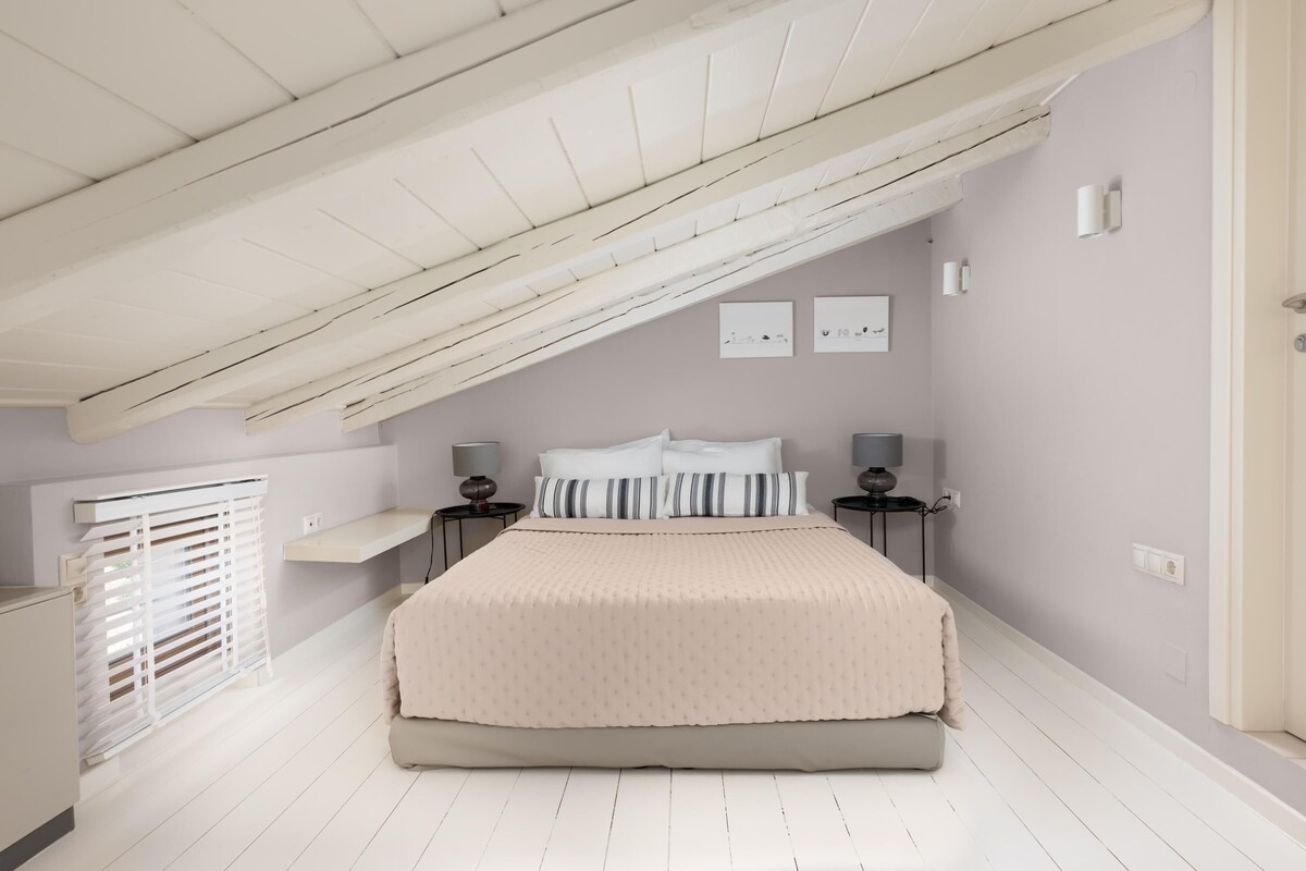 A bedroom with a sloped ceiling, a bed with a beige bedspread, two bedside tables with lamps, and a window with blinds.