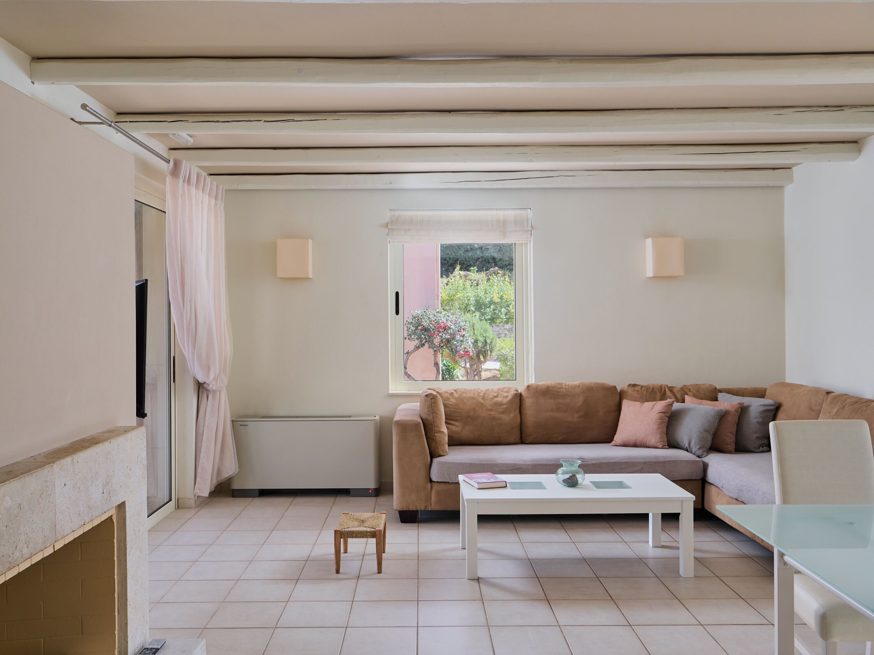 Una sala de estar con un sofá color beige, una mesa de centro blanca y un taburete pequeño de mimbre. Se ve una ventana con vistas a zonas verdes.