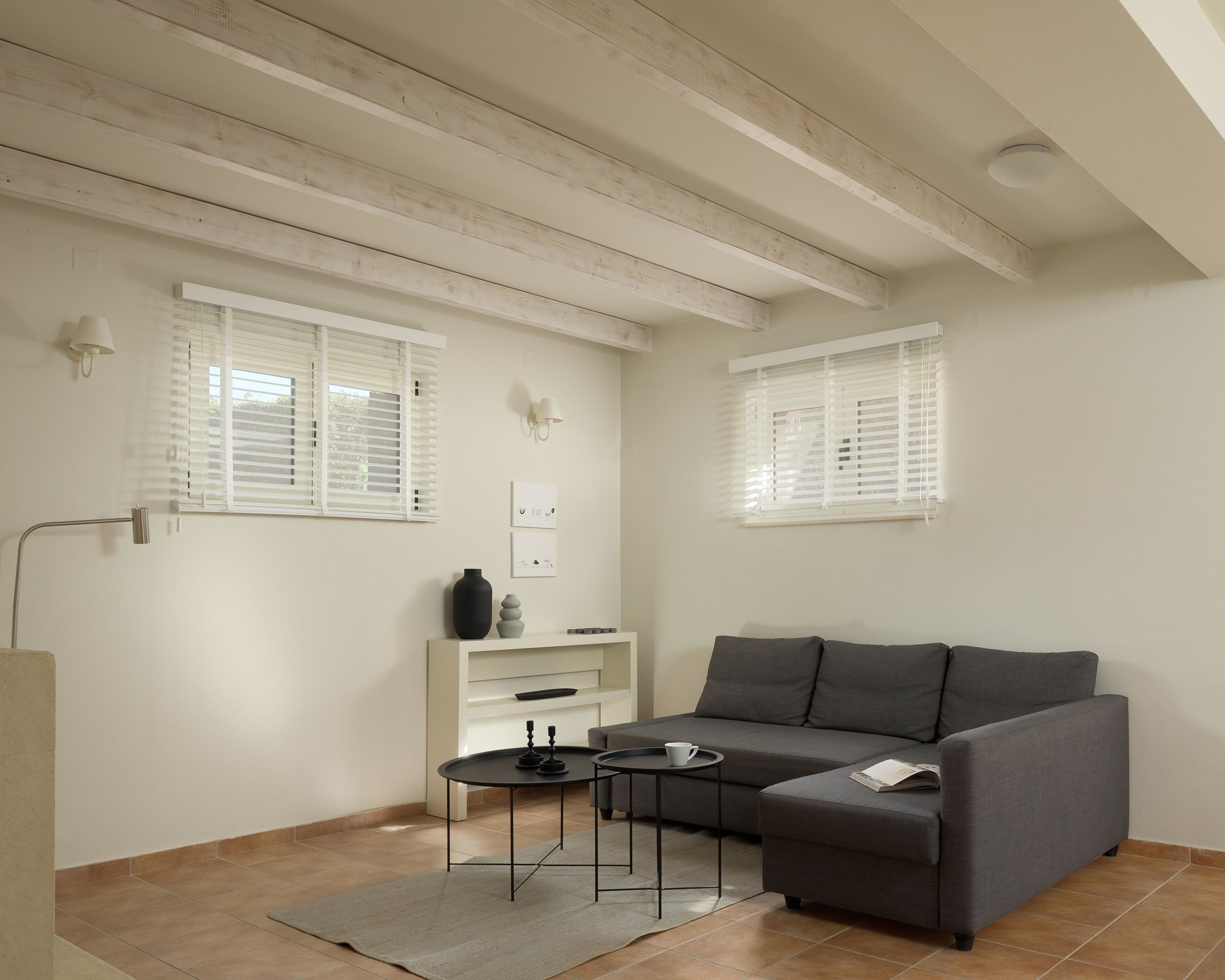 A living room with a grey sofa, a small side table, a white console table, and a floor lamp. There are two windows with blinds, a ceiling light, and a white wall.
