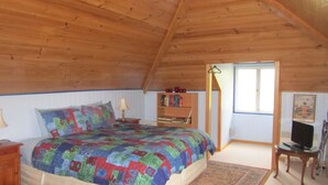 A bedroom with a wooden ceiling, a bed with a colorful quilt, a bedside table with a lamp, a bookshelf, a television, and a window. - Triabunna Cabin and Caravan Park (Triabunna)