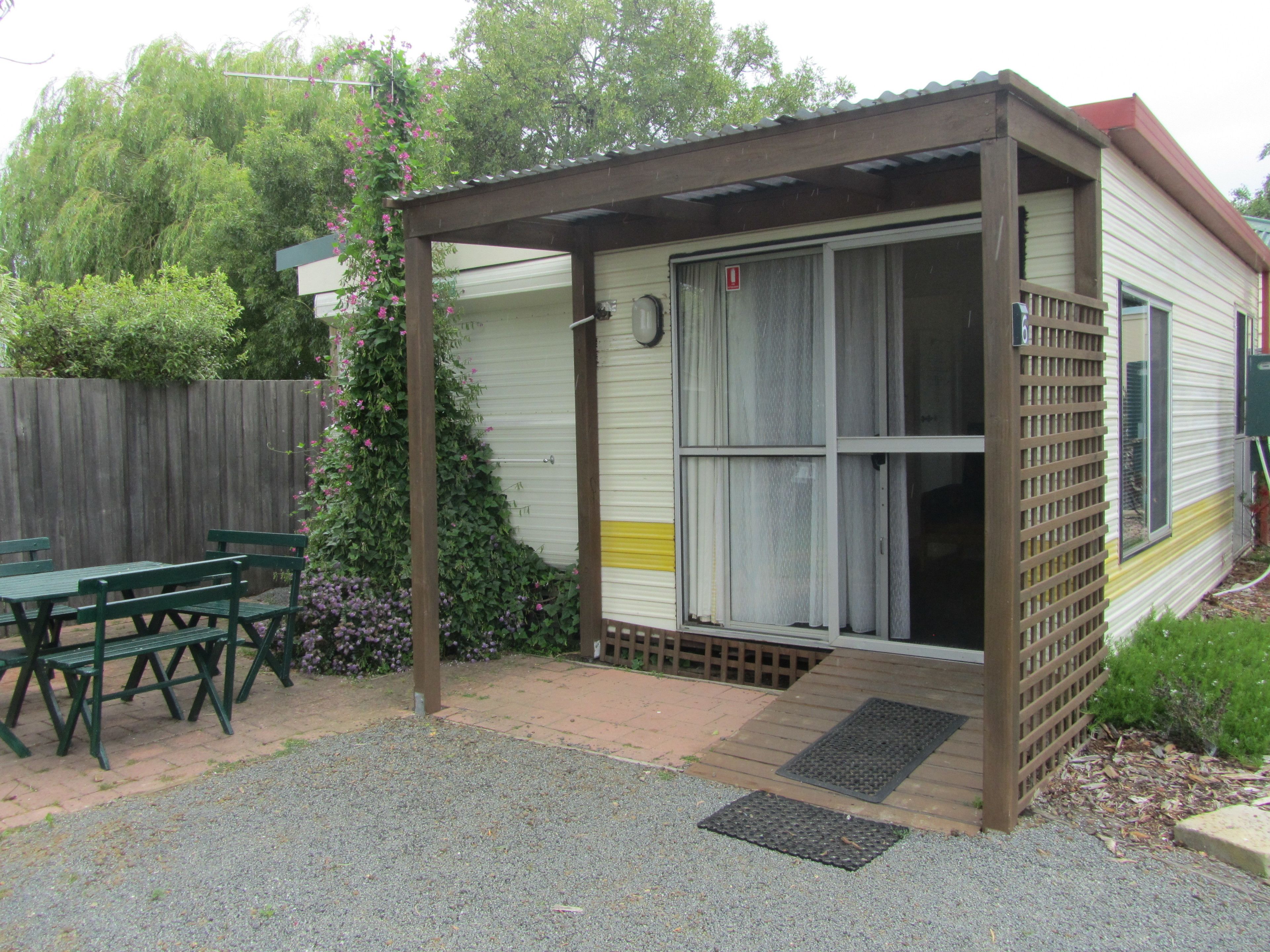 A small, single-story mobile home with a covered porch, a patio with a table and chairs, and a garden area with flowering plants.