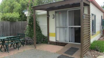A small, single-story mobile home with a covered porch, a patio with a table and chairs, and a garden area with flowering plants.