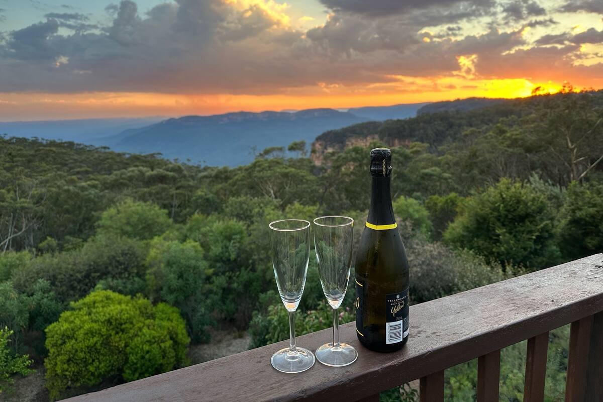 Una botella de Chardonnay Yellow Label y dos copas de vino vacías sobre una baranda de madera, con una vista panorámica de un bosque y montañas al atardecer.