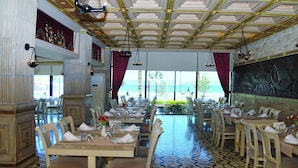 Un restaurant amb taules preparades per dinar, un terra amb dibuixos i vista al mar a través de grans finestres.