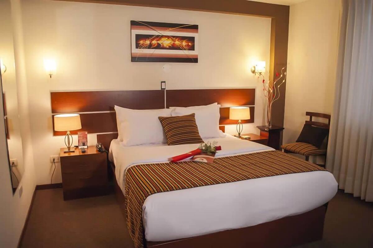 Une chambre d’hôtel avec un grand lit, deux tables de chevet avec des lampes, une tête de lit ornée d’un tableau et une chaise avec un coussin.