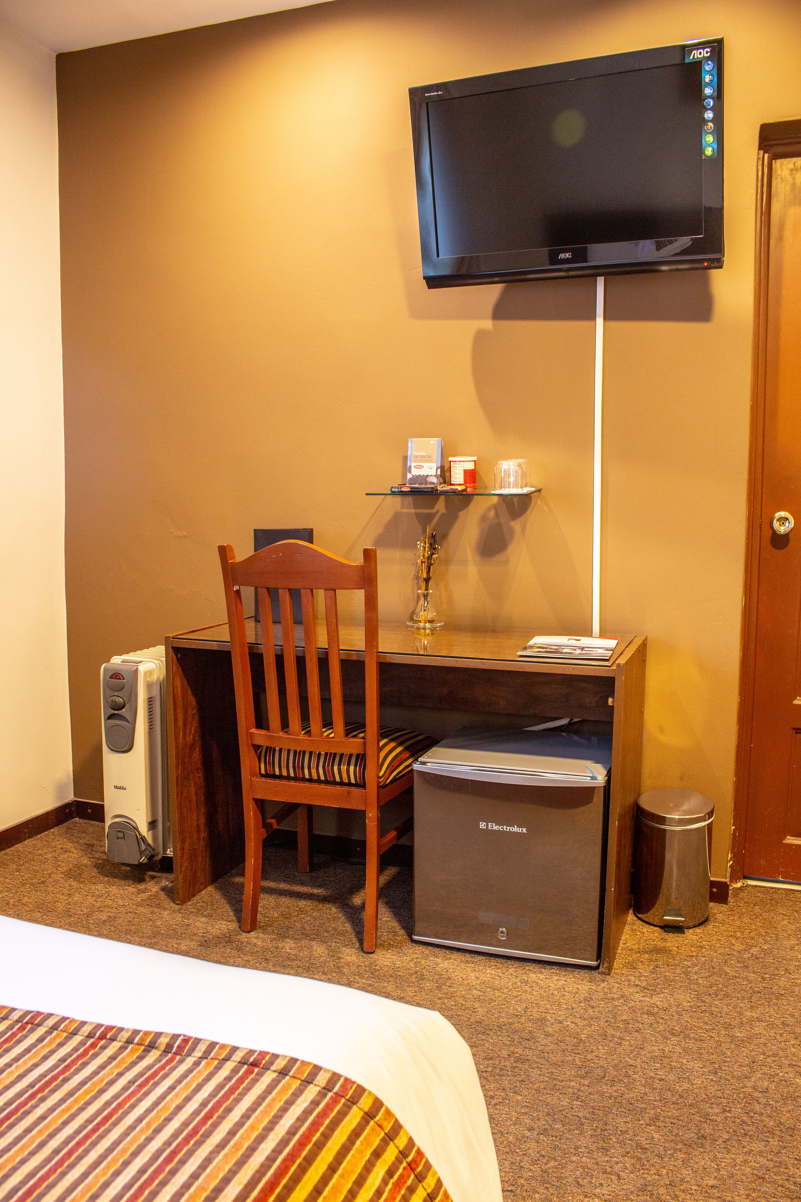 Habitación de hotel con cama, escritorio con silla, televisor montado en la pared, calefactor, cubo de basura y un refrigerador pequeño.