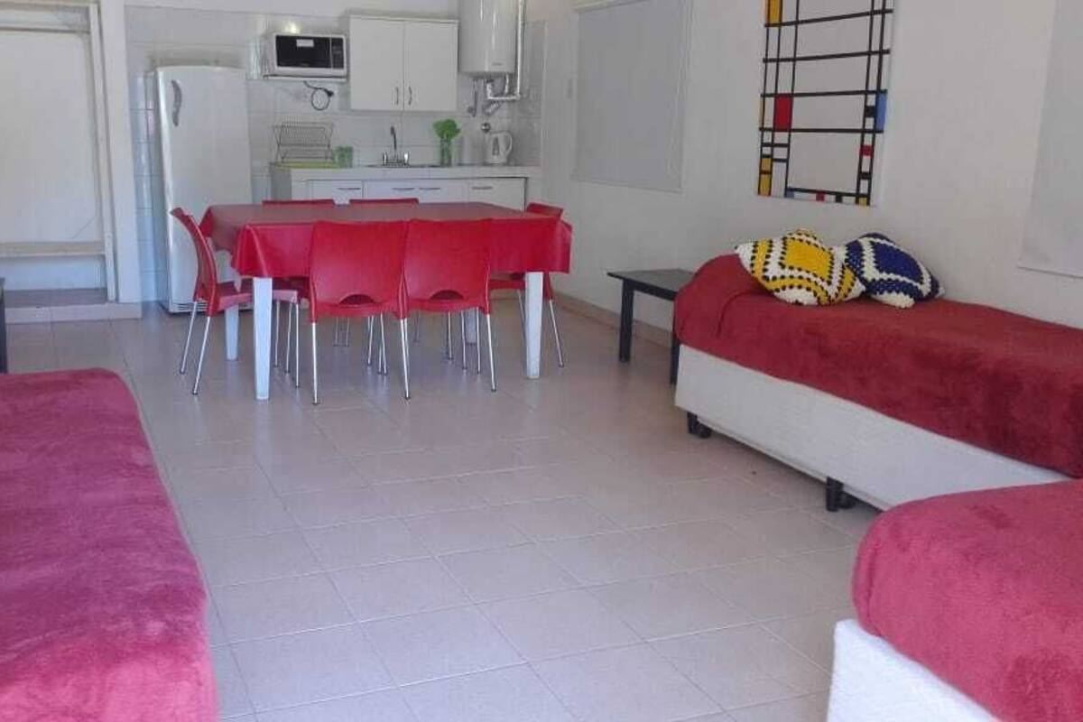 A living room with a red sofa, a dining table with red chairs, a kitchen area with white cabinets, and a colorful abstract art piece on the wall.