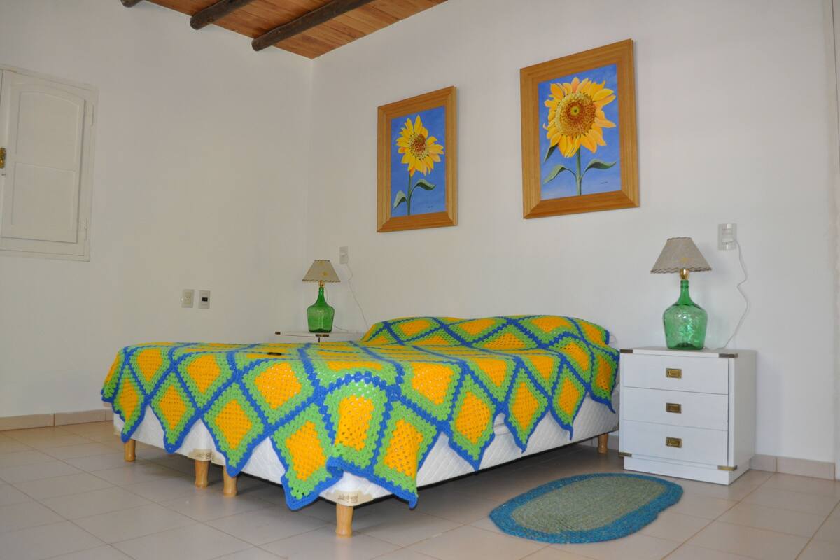 A bedroom with a bed covered in a colorful blanket, a nightstand with lamps, and two framed sunflowers on the wall.