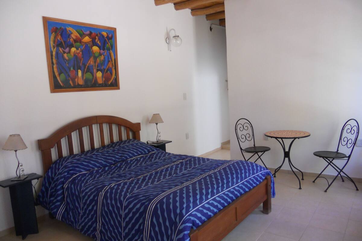 A bedroom with a bed, two nightstands, a table with two chairs, a painting on the wall, and a ceiling with wooden beams.