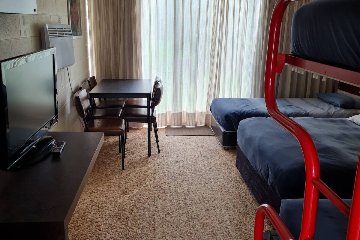 Una habitación con literas, un escritorio con computadora, una silla, un televisor y una ventana con cortinas.