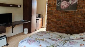 A bedroom with a bed, a desk, a television, a heater, and a brick wall with a floral painting.