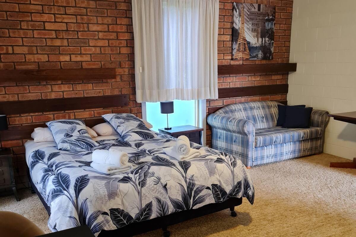 Un dormitorio con cama, sofá, escritorio, ventana con cortinas, ventilador de techo y pared de ladrillo.