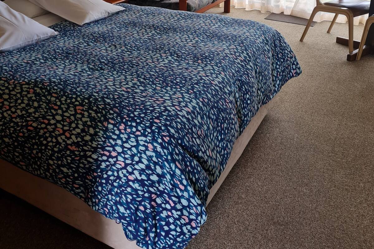 Habitación de hotel con una cama grande, un sofá, una silla y una mesita. Se ve una ventana con cortinas y un ventilador de techo.