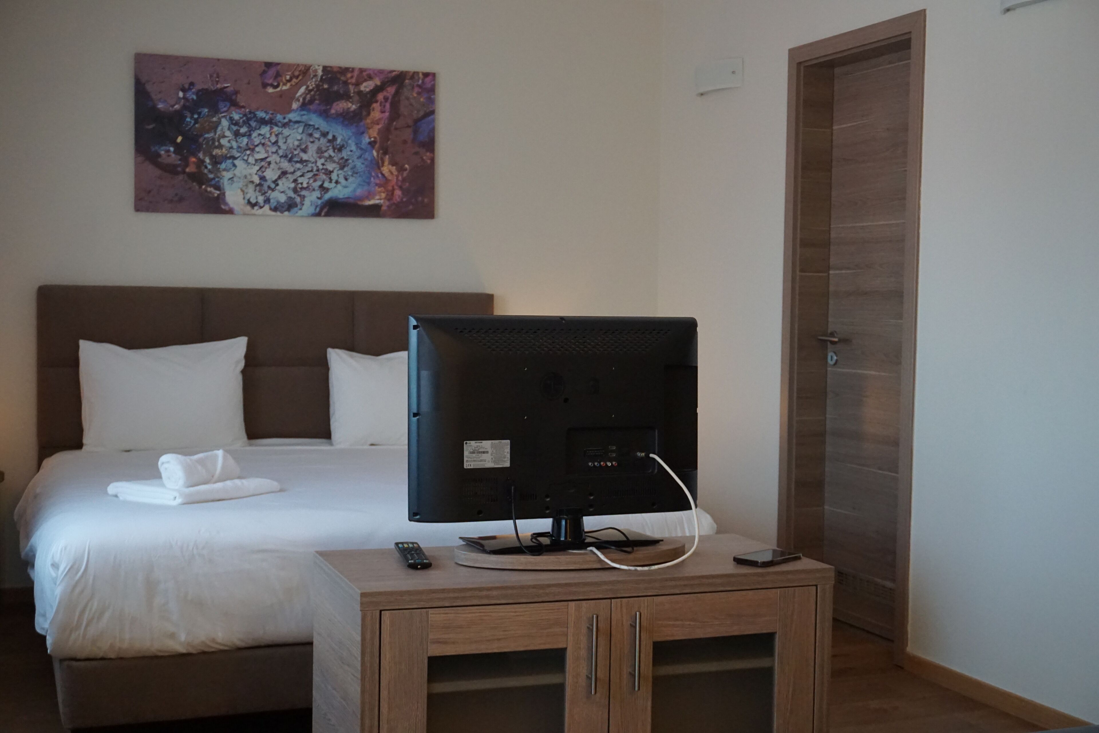 Habitación de hotel con una cama, un televisor sobre un soporte de madera, un control remoto y un cuadro en la pared.