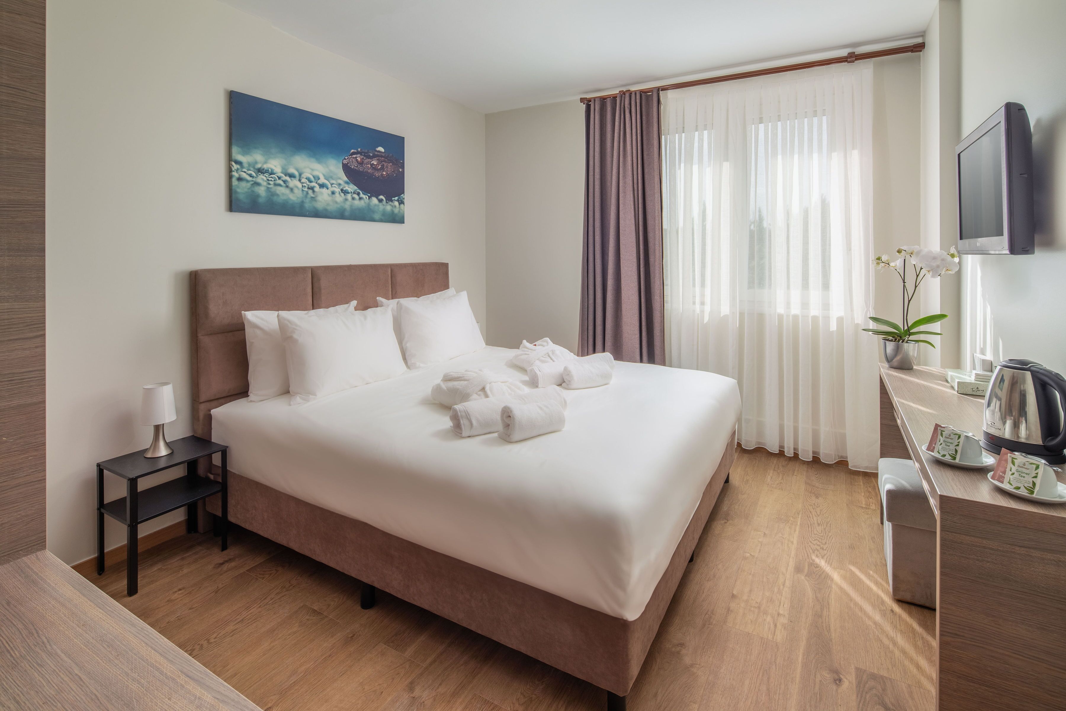 A neatly made bed with white linens and towels, a bedside table with a lamp, a television on the wall, and a window with sheer curtains.