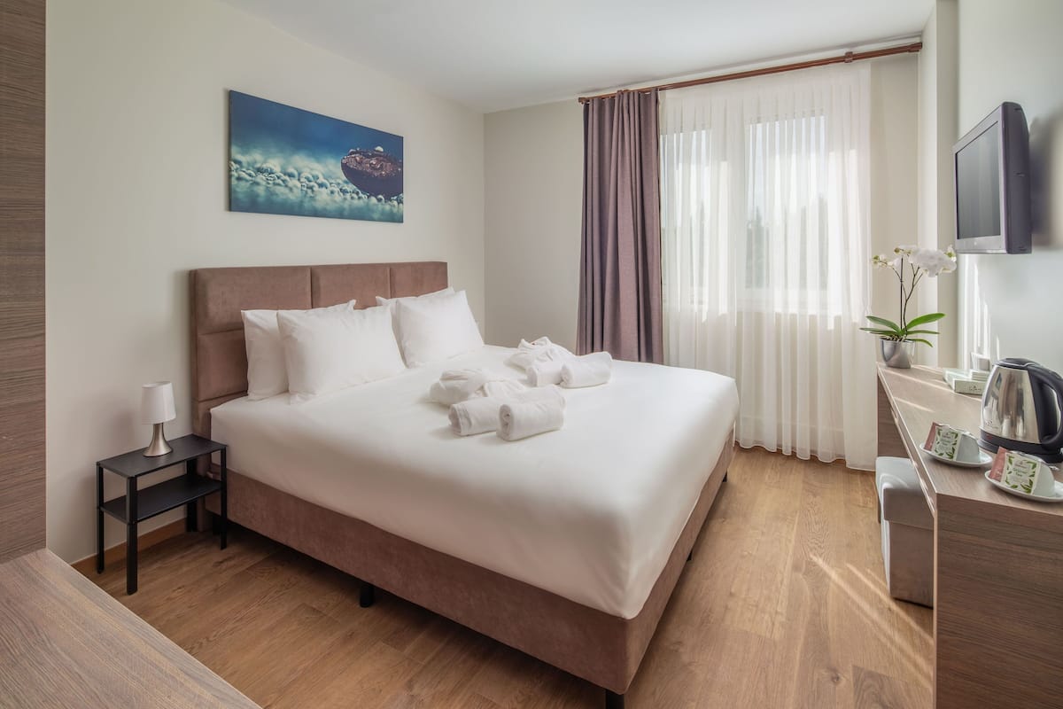Una cama bien tendida con sábanas y toallas blancas, una mesita de noche con una lámpara, un televisor montado en la pared y una ventana con cortinas traslúcidas.
