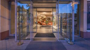 Ein Hotel mit Eingang, einer Glastür, einem Empfangsbereich mit orangefarbenen Sesseln und einem einsehbaren Innenbereich, einschließlich einem Essbereich.