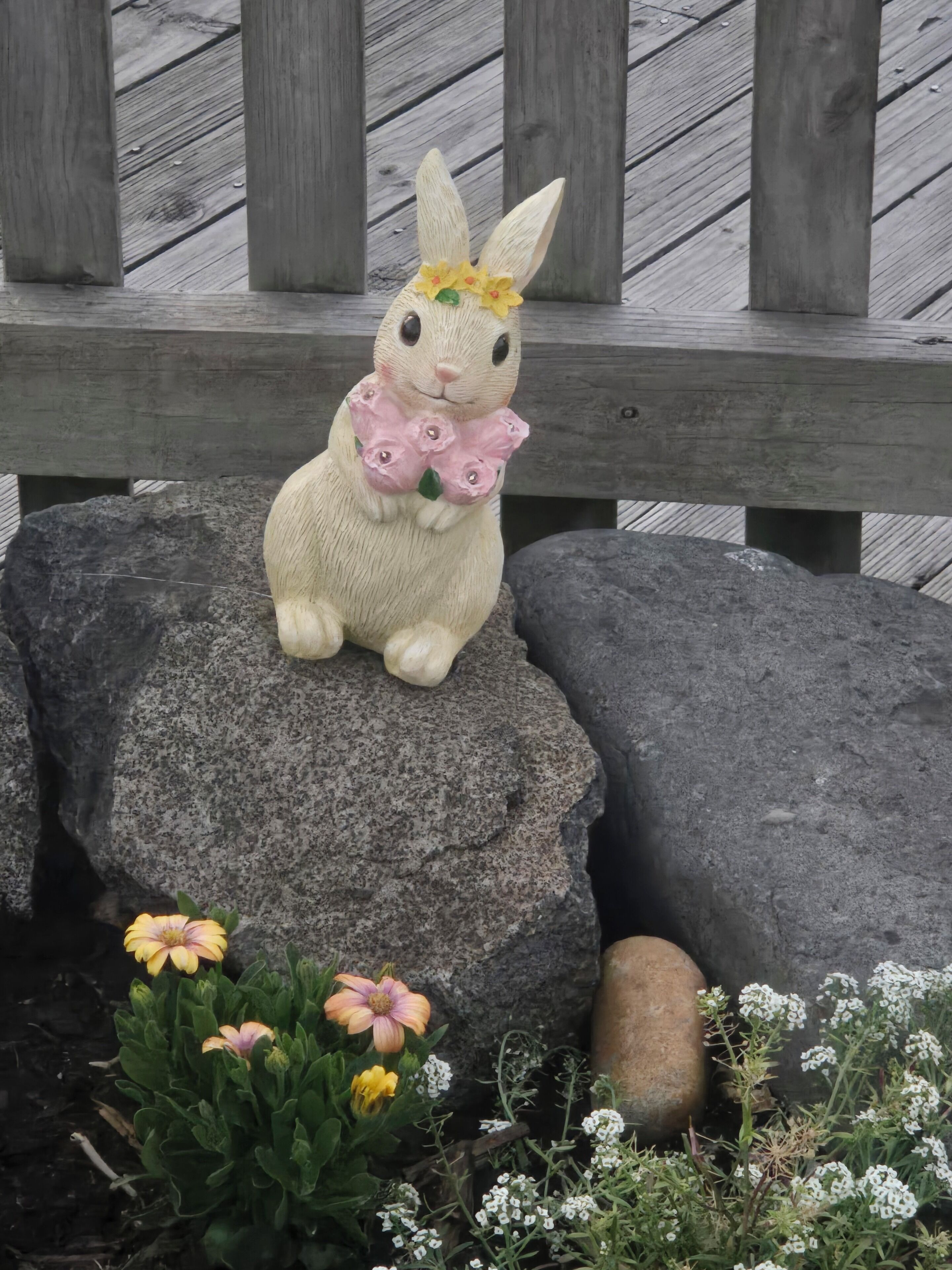 A ceramic rabbit figurine on a rock, surrounded by flowers and a wooden bench.