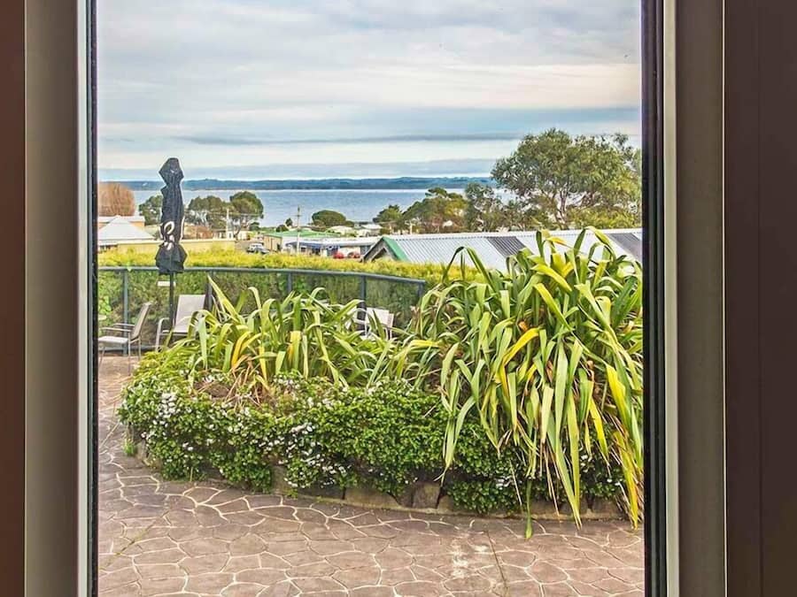 A view from a balcony with a stone floor, lush greenery, and a distant body of water.