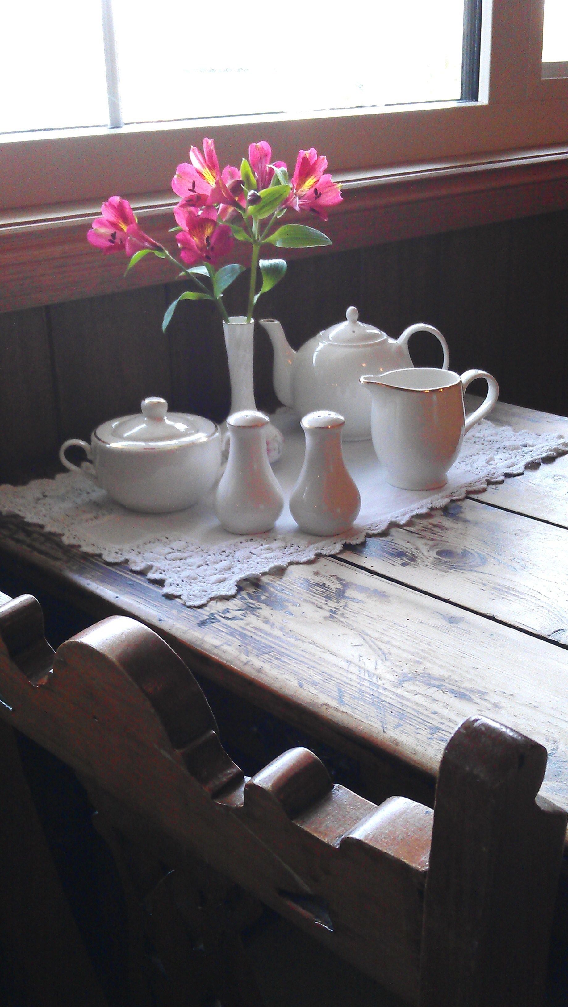 Une table en bois surmontée d’un chemin de table en dentelle, une théière blanche, deux sucriers blancs et une tasse blanche.
