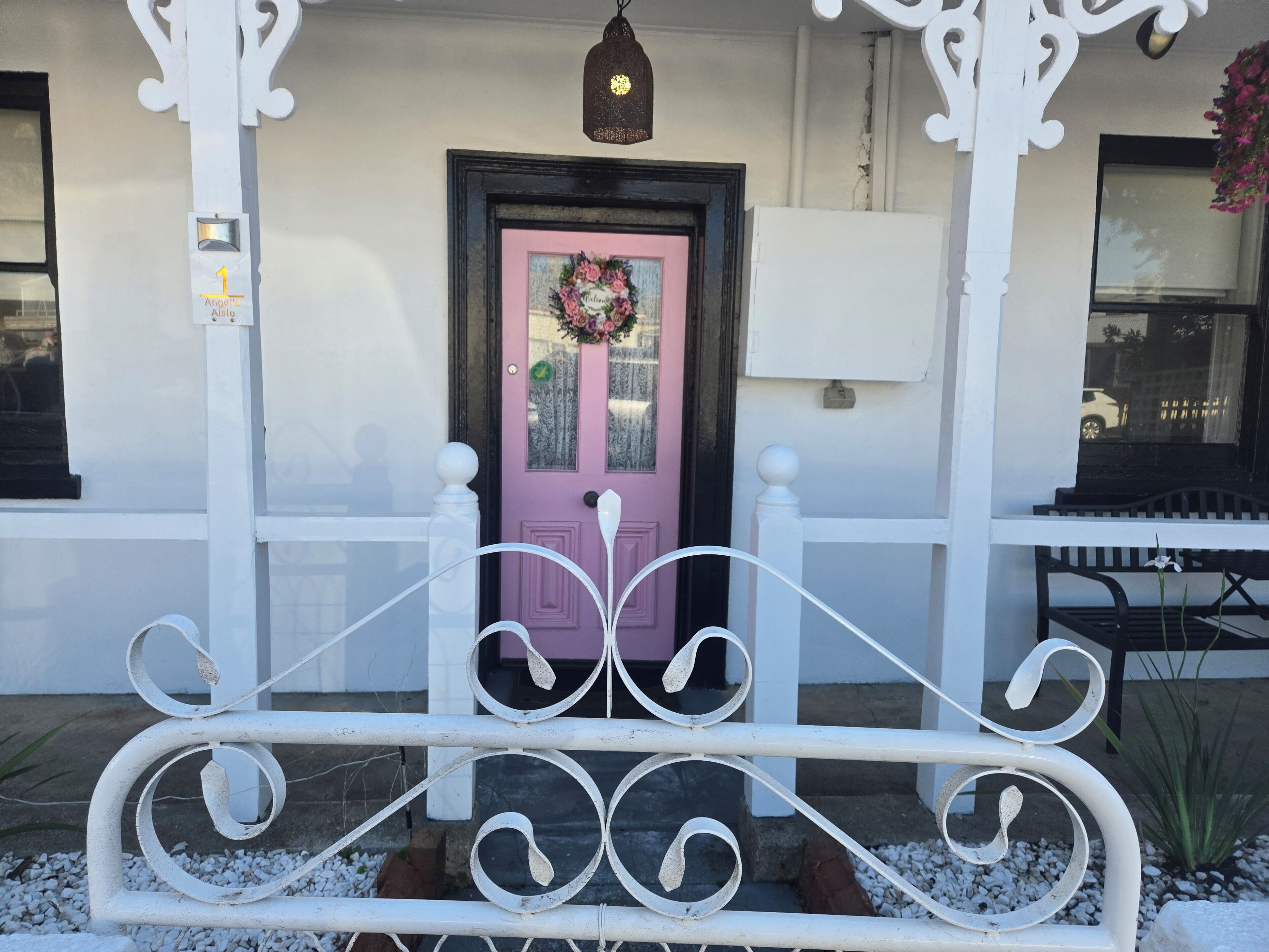 Un banco de metal blanco frente a una puerta rosa adornada con un adorno, una valla de piquetes blancos y un farol colgante.