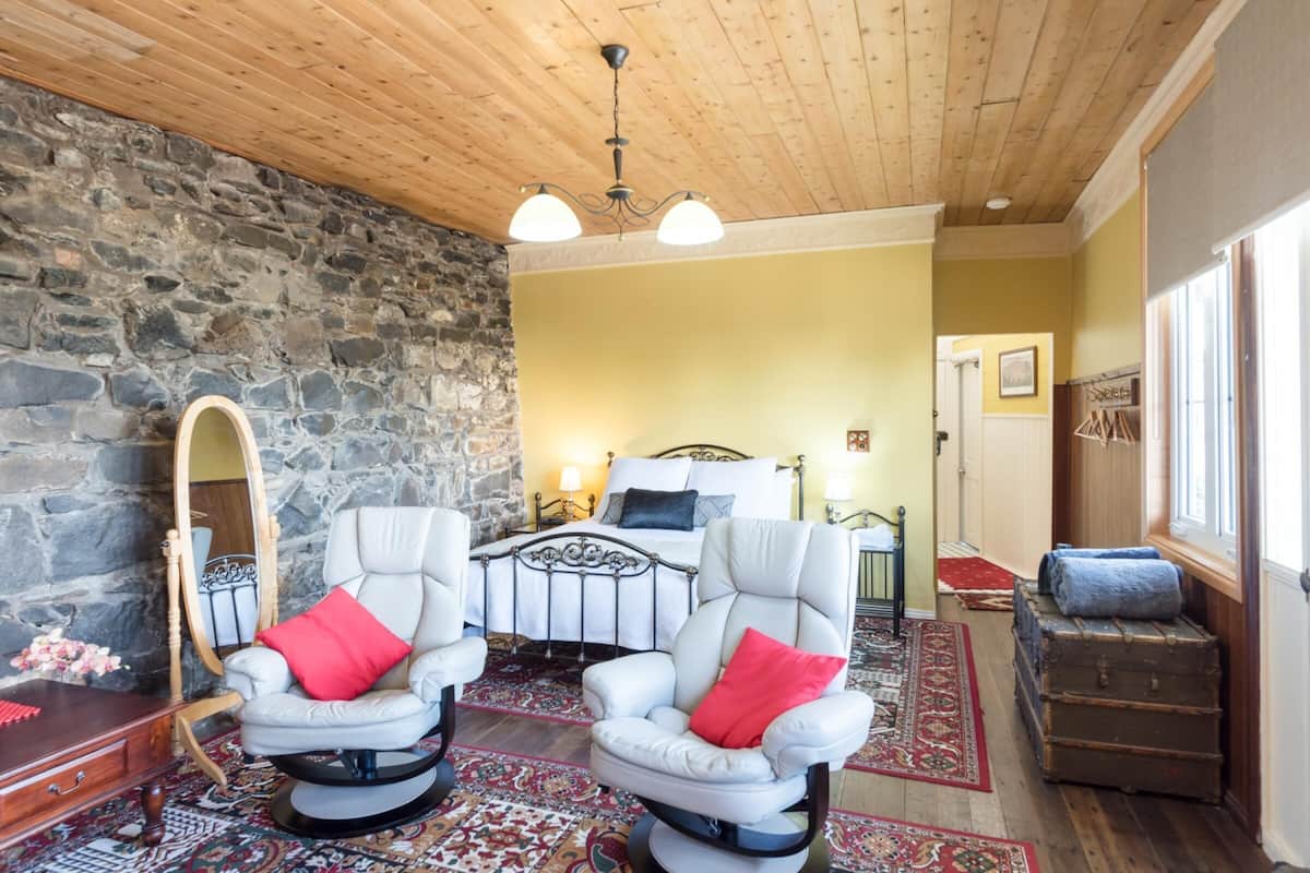 A cozy room with a stone wall, a bed with a metal frame, two white armchairs with red cushions, a wooden ceiling, and a large mirror.