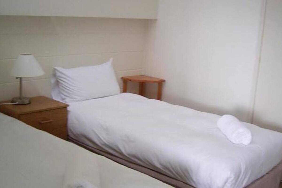 A single bed with a white mattress and pillow, a bedside table with a lamp, and a wooden nightstand.